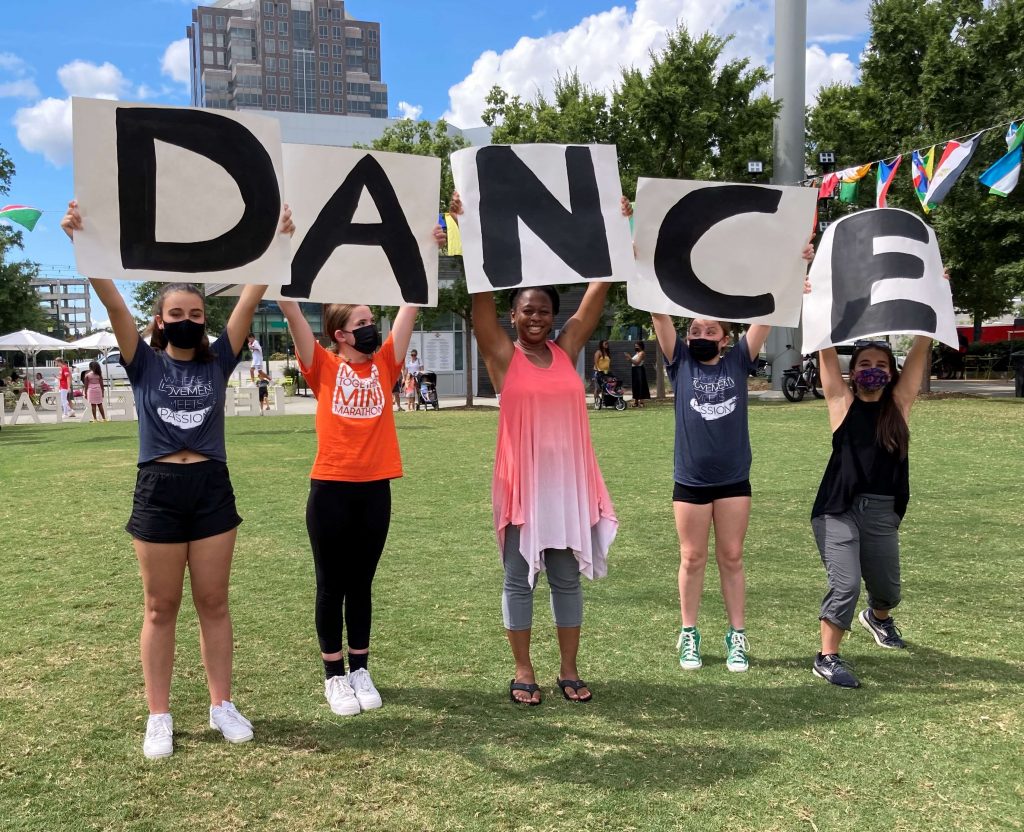 Five dancers in a sunny park setting standing in a row, holding up signs spelling the word dance.