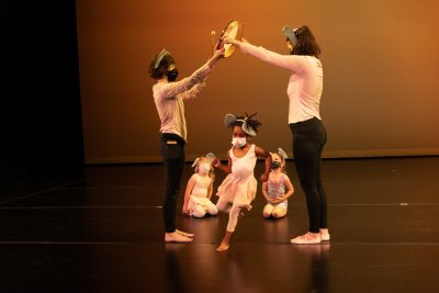Two dance teachers on a stage form an archway with their arms while a young toddler dancer runs between them.