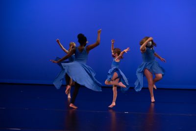 A group of four young dancers in a circle, wearing matching blue dresses, performing a lively dance on a stage.