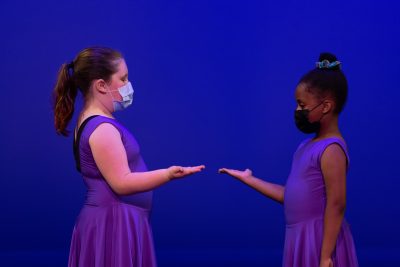Two Mini Performance Company Dancers wearing purple dresses performing a duet on stage.