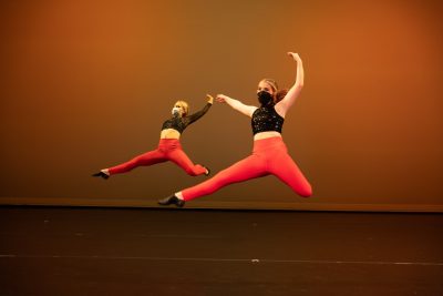 Two Teen Performance Company students mid-leap while performing on stage.