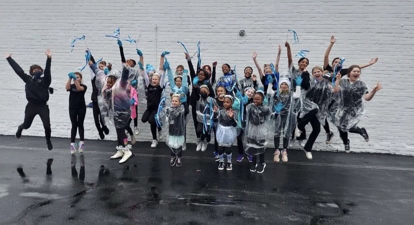 A large group of Dance Project students in front of a white brick wall jumping in the air for a group photo.
