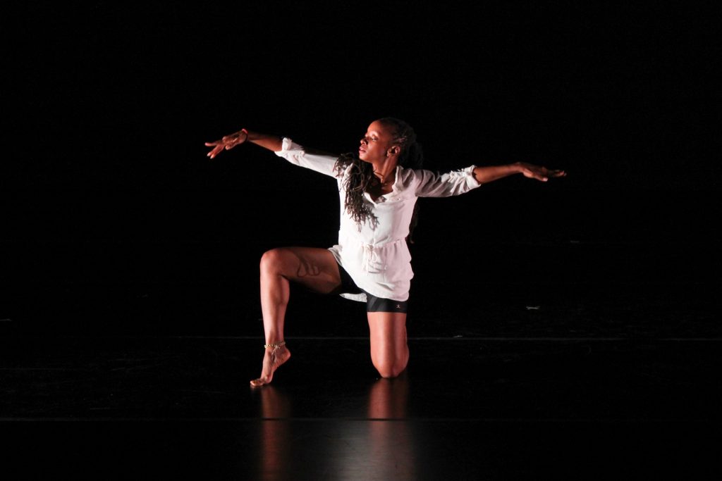 A dancer poses on a dark stage, kneeling with one leg out to the side and both arms outstretched to each side.