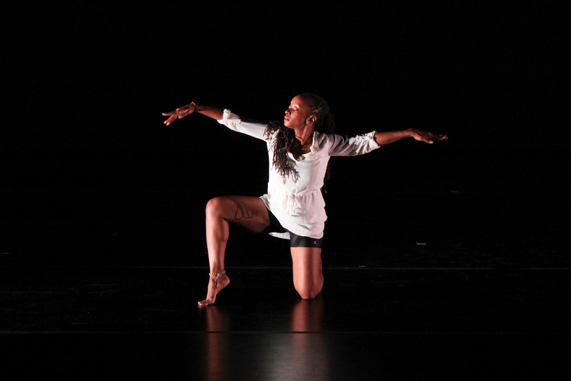 A dancer poses on a dark stage, kneeling with one leg out to the side and both arms outstretched to each side.