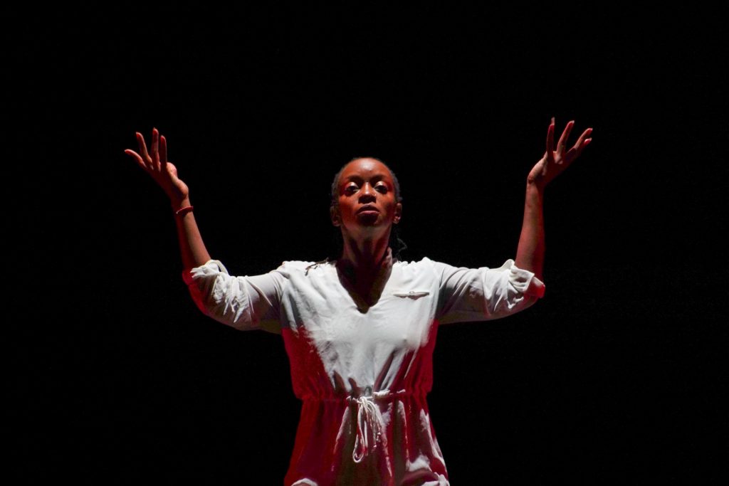 A dancer against a dark stage backdrop, lit from above, facing forward with both hands raised either side of her head.