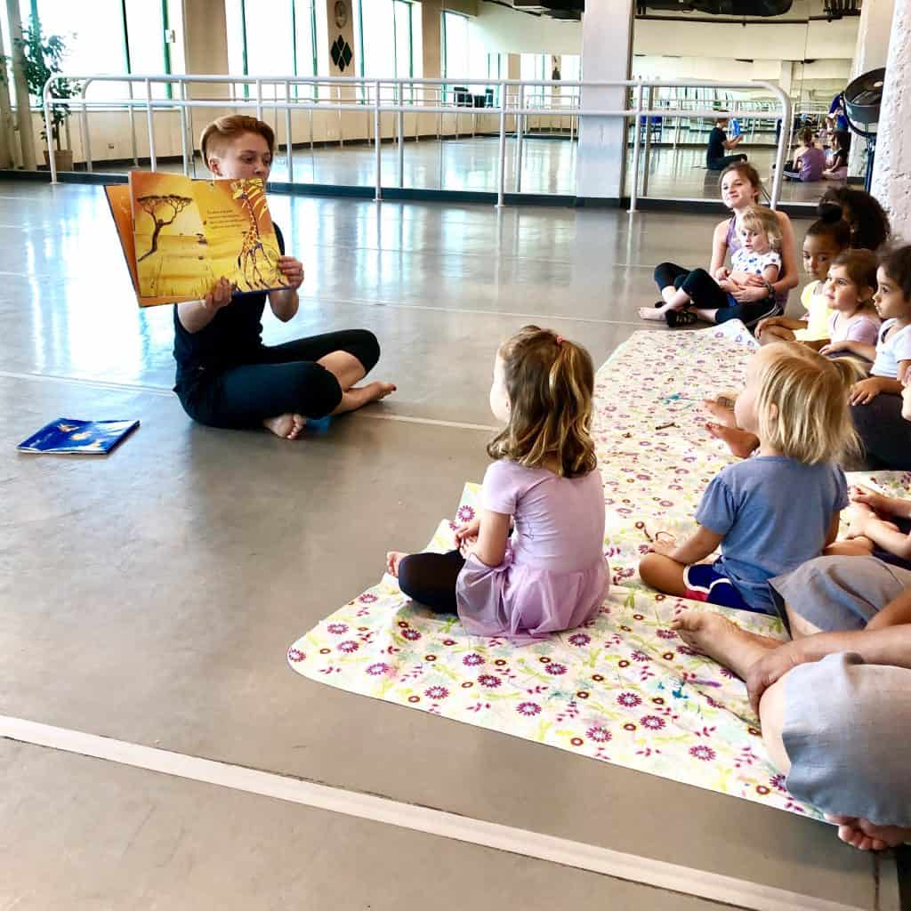 Children sitting in a dance studio listening to a story being read.