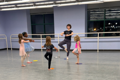 Instructor Magalli Morana leads a small group of young dancers in a circle, all standing with one leg lifted in a passe position.