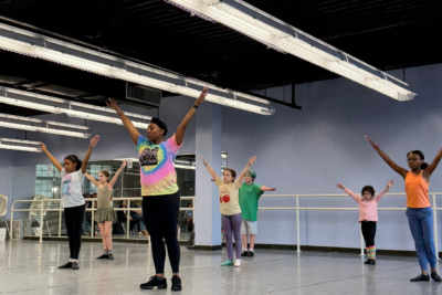 Instructor Milanda leads a youth jazz class, standing with her arms raised in a V position.
