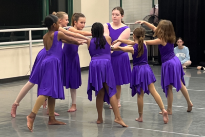 A group of 7 tween dancers in bright purple dresses pose in a circle with their hands meeting in the middle.