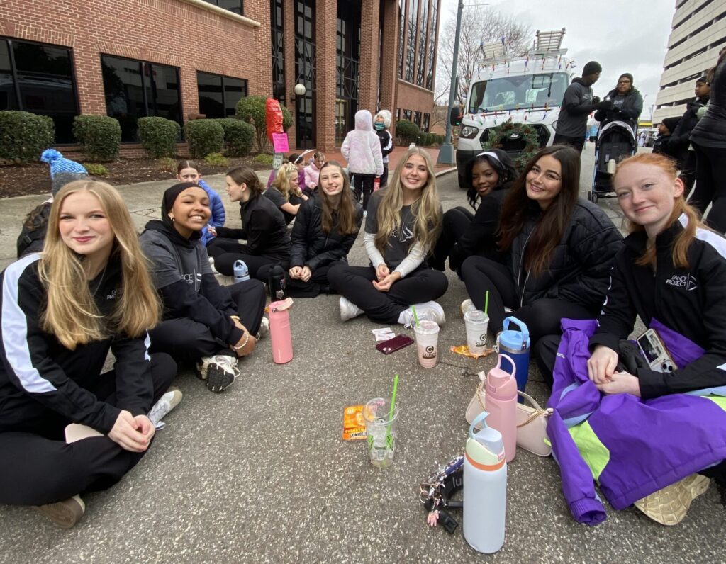 A group of teenaged Dance Project dancers sits on the street waiting for the Holiday Parade to begin.