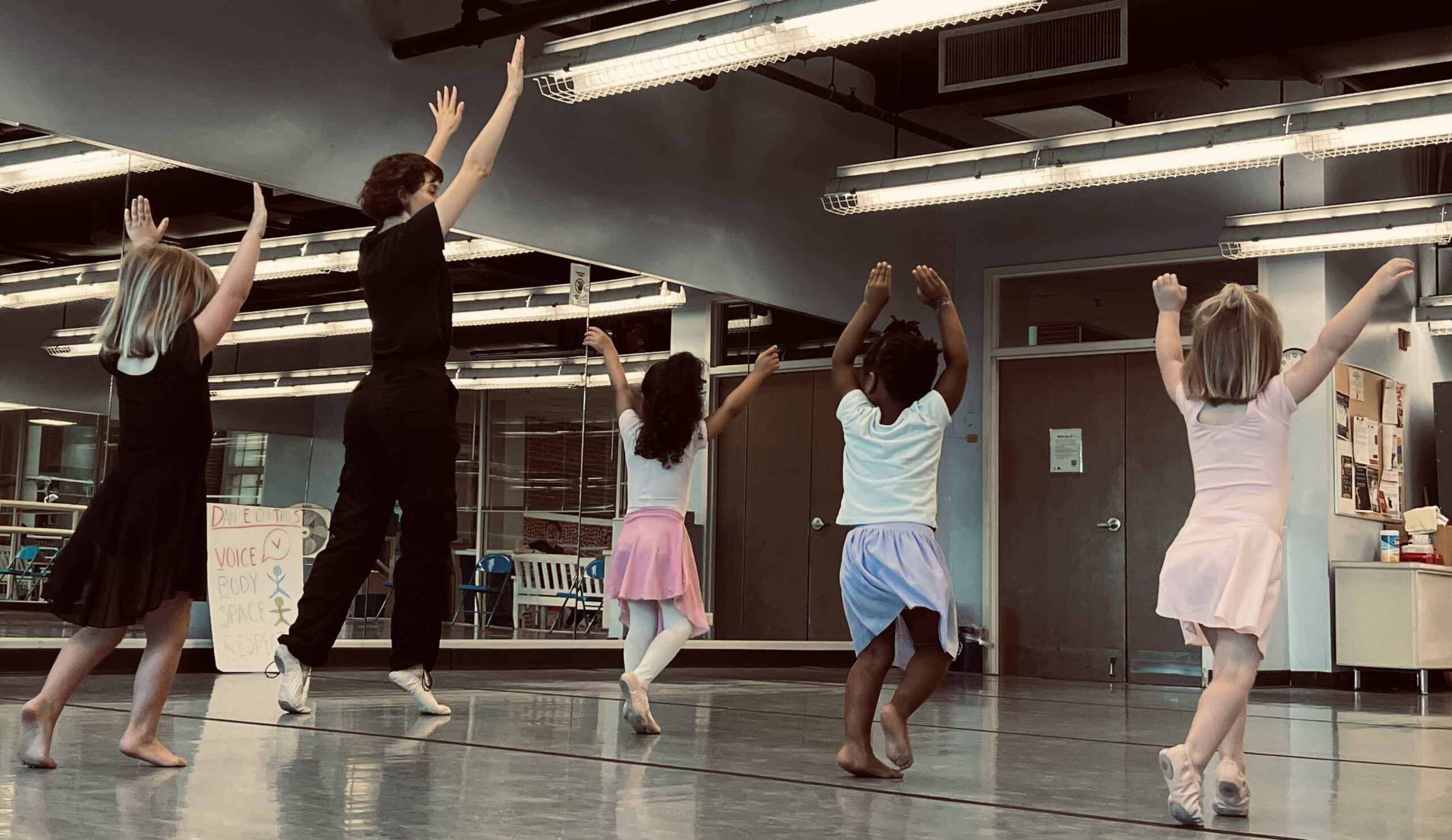 A dance teacher reaches both hands up to the sky and 4 pre-school aged dancers imitate her