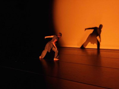 Dancer Alyah Baker in NC Dance Festival performance, her brown tunic and pants glowing in the gold spotlight that casts her shadow on the cyc at the back of the stage.