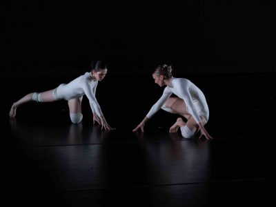 Two white women in white unitards point to a spot on the dance floor in Eric Mullis's choreography.
