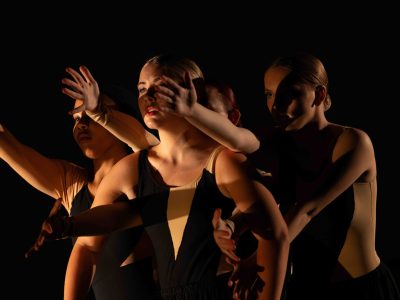 A cluster of young women in black and nude costumes make a dramatic pose, with their arms reaching through the arms and past the shoulders of the dancer in the front.
