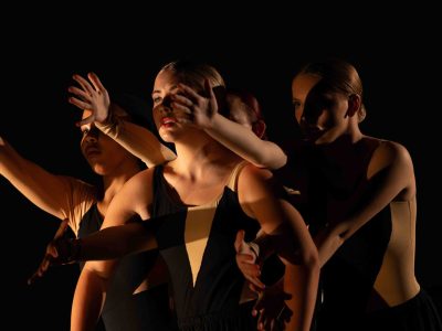 A cluster of young women in black and nude costumes make a dramatic pose, with their arms reaching through the arms and past the shoulders of the dancer in the front.