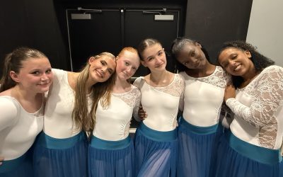 A group of 6 high school senior dancers in white leotards and teal skirts pose with their arms around each other during Dance Project's Spring Showcase.