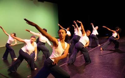 A group of adults ranging from 20s to 50s dances on a stage, lunging on one leg while extending their top arm on a diagonal.