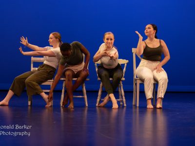 Four dancers strike different dance poses while seated on chairs on a stage.