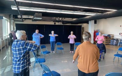 A group of older adults stand with their arms in a round shape in front of them during Dance Project's class for Parkinson's.