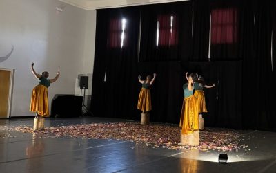 Four dancers stand on top of short tree stumps, arranged in a square in a dance studio theater. They wear bright gold skirts and the ground between the stumps is covered with silk flower petals.
