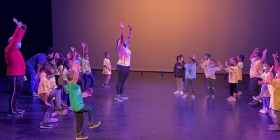 A large group of visiting elementary school students dancing together with Dance Project teacher, Vania Claiborne, on a stage.