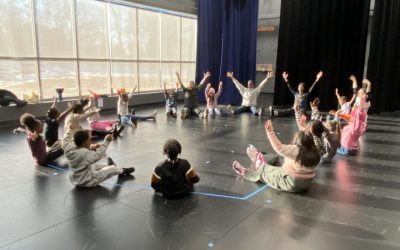 20 first grade students at Peck Elementary sit on the school stage with their hands raised during their dance class warm up.