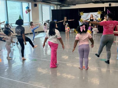 A large group of young dancers stand in a circle with one leg extended into the middle during Dance Project's Birthday Bash class.